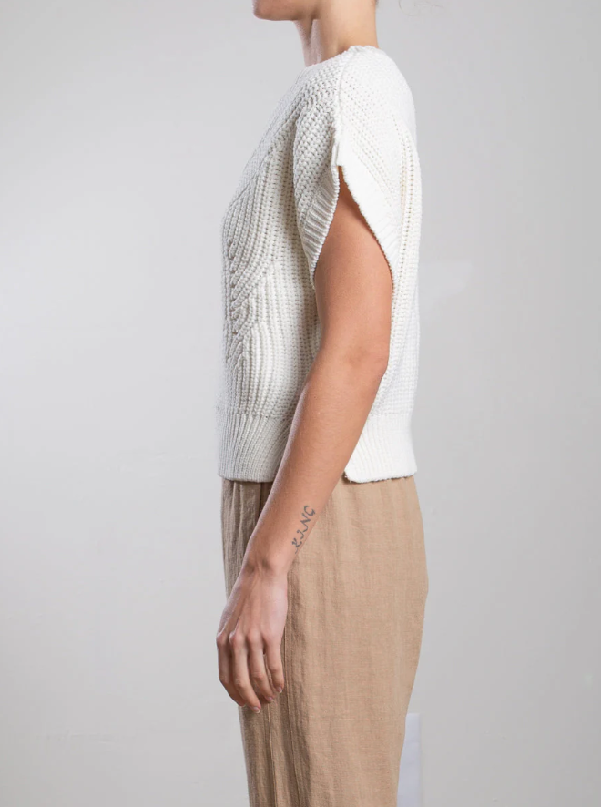 Facing left, the person wears a Pharaoh Edie Shell sweater in white with ribbed accents and beige pants. Their arm displays a "LOVE" tattoo against a plain, neutral background.