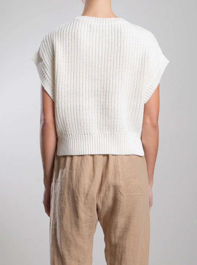 The individual wears a Pharaoh Edie Shell, featuring ribbing accents with its short-sleeved white knit design, ideally paired with lightweight, high-waisted beige pants. The scene unfolds against a simple light-colored wall.