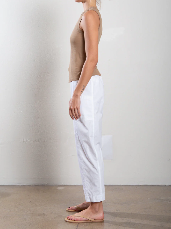 A person stands barefoot on a concrete floor, facing a plain white wall, wearing Pharaoh's beige sleeveless top paired with the Torrance Pant—garment-dyed barrel-leg pants featuring an elasticized waist.