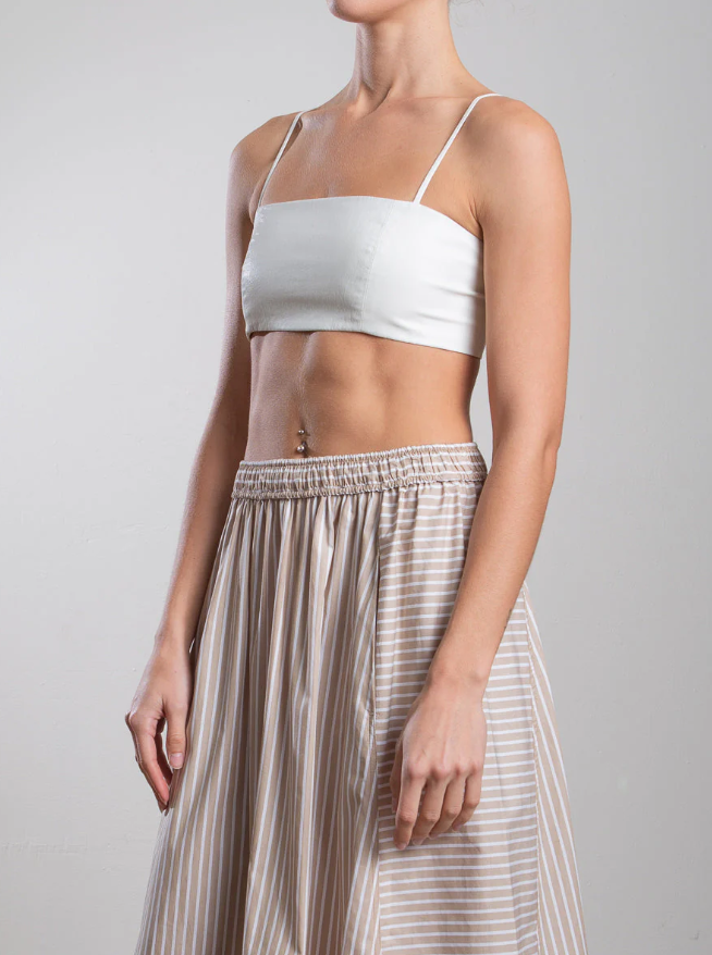 Sporting a Toga Bralette by Pharaoh, a person in a white crop top pairs it with a high-waisted beige skirt featuring white stripes. The ensemble showcases their midriff, with their left arm slightly bent, exuding bralette-inspired flair against a plain background.