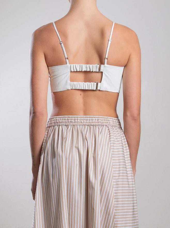 The image showcases a person from behind wearing Pharaoh's Toga Bralette, a white strapless piece with adjustable straps and a rectangular cutout, paired with a high-waisted beige and white vertically striped skirt. The plain light-colored background highlights the outfit's elegance.