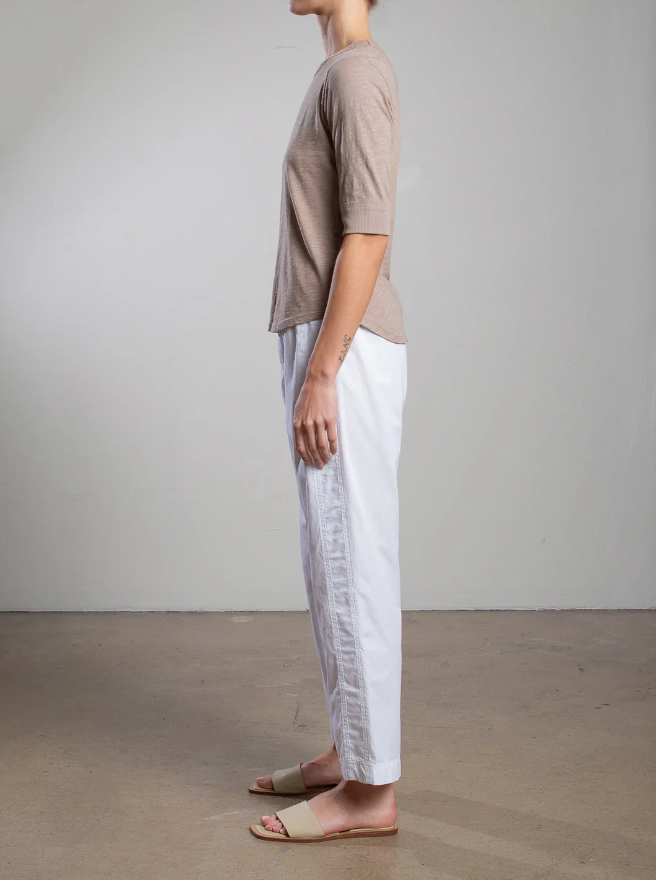 In a minimalist room with a light-colored wall and concrete floor, someone stands sideways wearing Pharaoh's Andy Pant in white, featuring side stripes and an elastic waist, paired with a beige T-shirt and beige sandals.