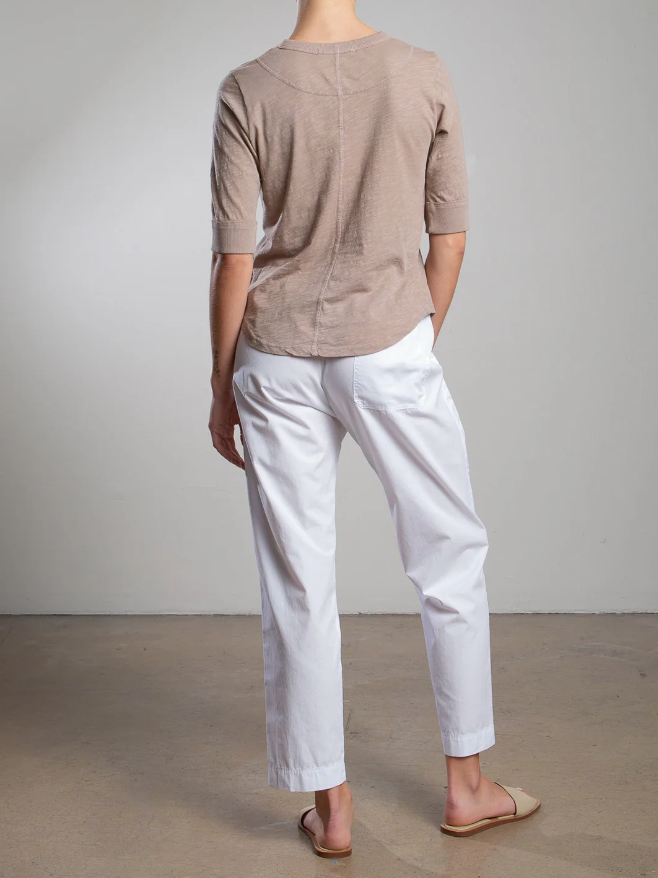 Rear view of a person on a concrete floor, wearing a beige 3/4 sleeve top, Pharaoh's Andy Pant with an elastic waist, and sandals, all against a plain gray wall in a minimalist setting.