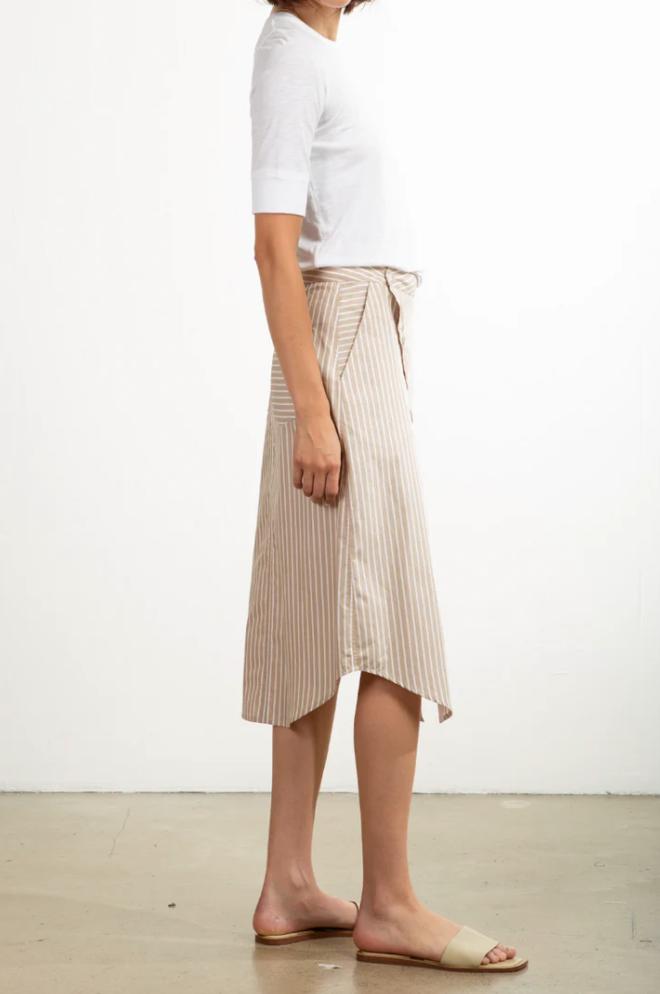 A person is standing in profile against a plain background, wearing a white short-sleeve top and the Pharaoh Nico Skirt with beige and white stripes, made in Los Angeles. They complete their look with beige slide sandals.