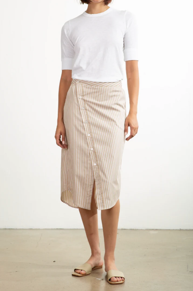 A person stands against a plain background wearing a 100% cotton white T-shirt with Pharaoh's beige striped Nico Skirt featuring an asymmetric placket, completed by chic beige slide sandals.