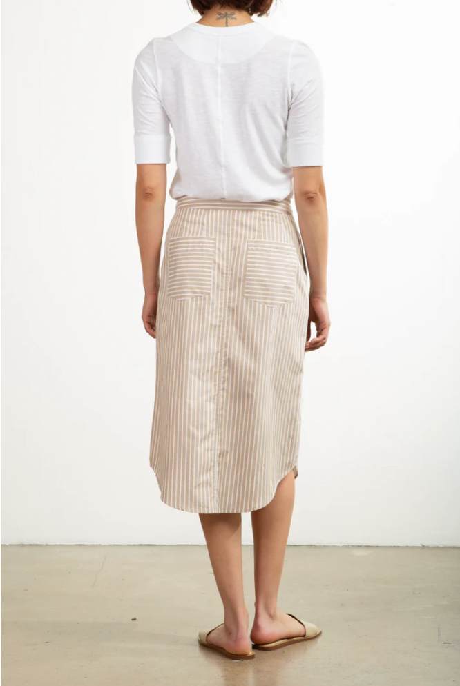 A person is seen from behind wearing a white shirt and the Pharaoh Nico Skirt—a knee-length beige asymmetric placket skirt with vertical stripes and two back pockets. Paired with beige flat sandals, this chic outfit stands out against an off-white wall background. Proudly made in Los Angeles.