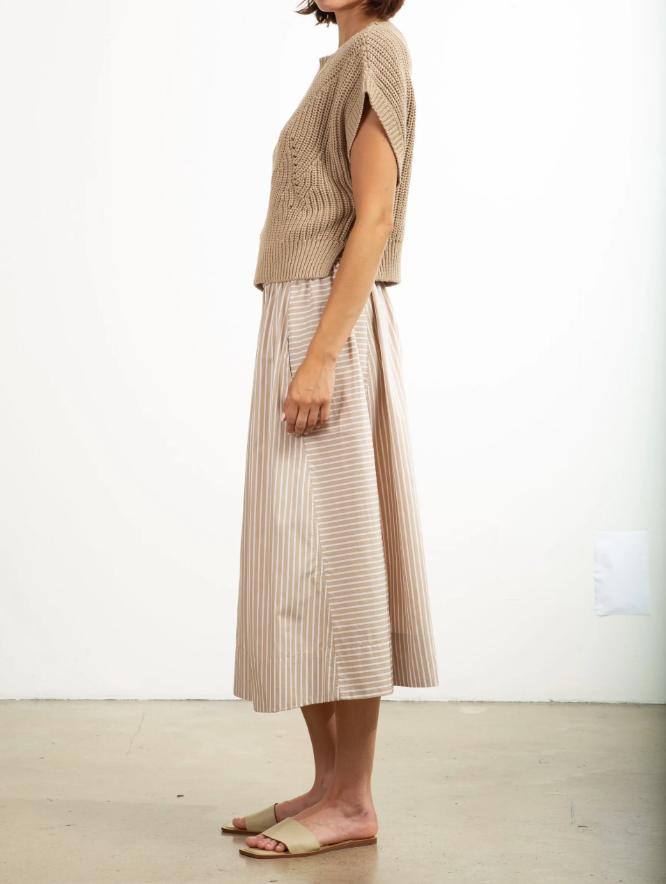 A side view shows a person in a beige short-sleeve top and the Pharaoh Claudia Skirt, a knee-length beige and white striped midi with an elastic waist. The ensemble is 100% cotton for comfort. They stand on a concrete floor in tan sandals against a plain white wall.