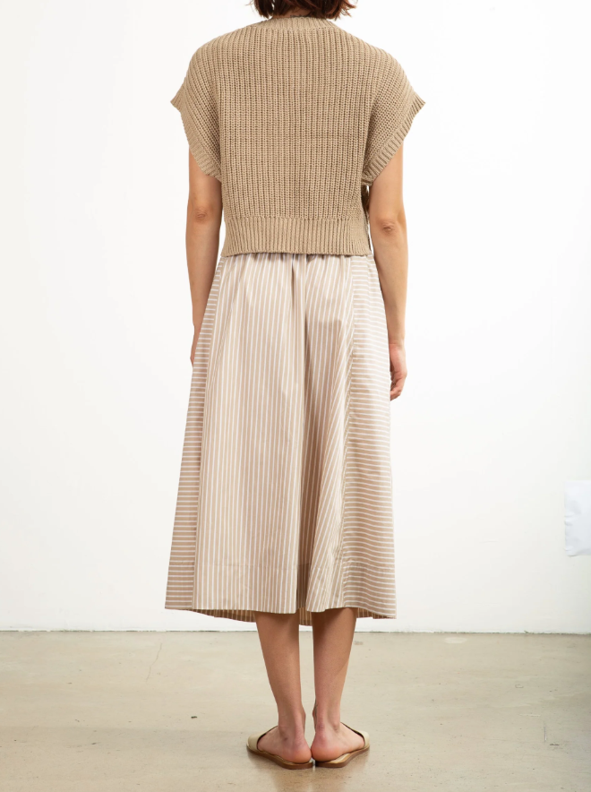 A person wears the Pharaoh Claudia Skirt, a beige and white striped 100% cotton midi with side pockets, paired with a brown knitted top and brown sandals. They stand on a light floor against a white backdrop.