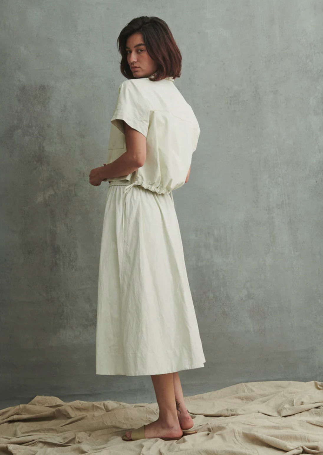 A woman with shoulder-length brown hair stands barefoot on a crumpled sheet, wearing the Claudia Skirt by Pharaoh. The light dress features a full midi skirt and an elastic waist, set against a textured gray wall for a minimalist setting. Made in Los Angeles.