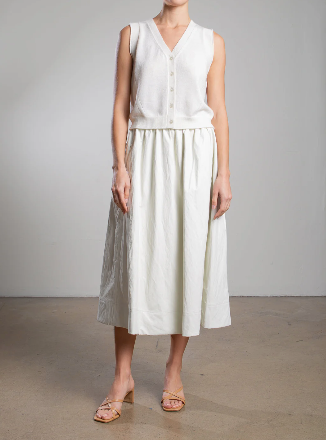 Against a plain backdrop, a person models Pharaoh's Claudia Skirt—a sleeveless white top pairs with it perfectly. Their beige heeled sandals enhance the ensemble, but their face remains unseen.