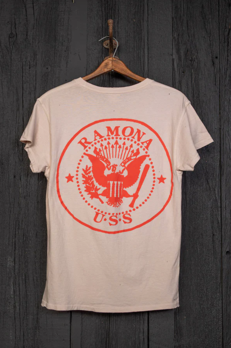 A Ramones T-shirt by Made Worn hangs on a wooden wall, showcasing a vintage unisex design. It features a large red eagle graphic with arrows and branch, encircled by "RAMONA" and "U*S*S". Handmade in Los Angeles, it has a beige color with a pink tea stain finish.