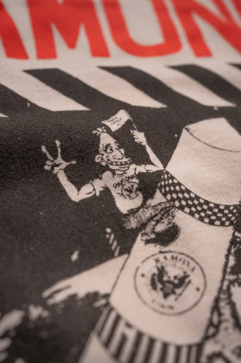 A black and white illustration by Made Worn features a cartoon character in a pointed hat, smiling and waving. It stands near an eagle-emblem cylinder, with a pink tea stain adding nostalgia. The word "RAMONES" is partially visible.