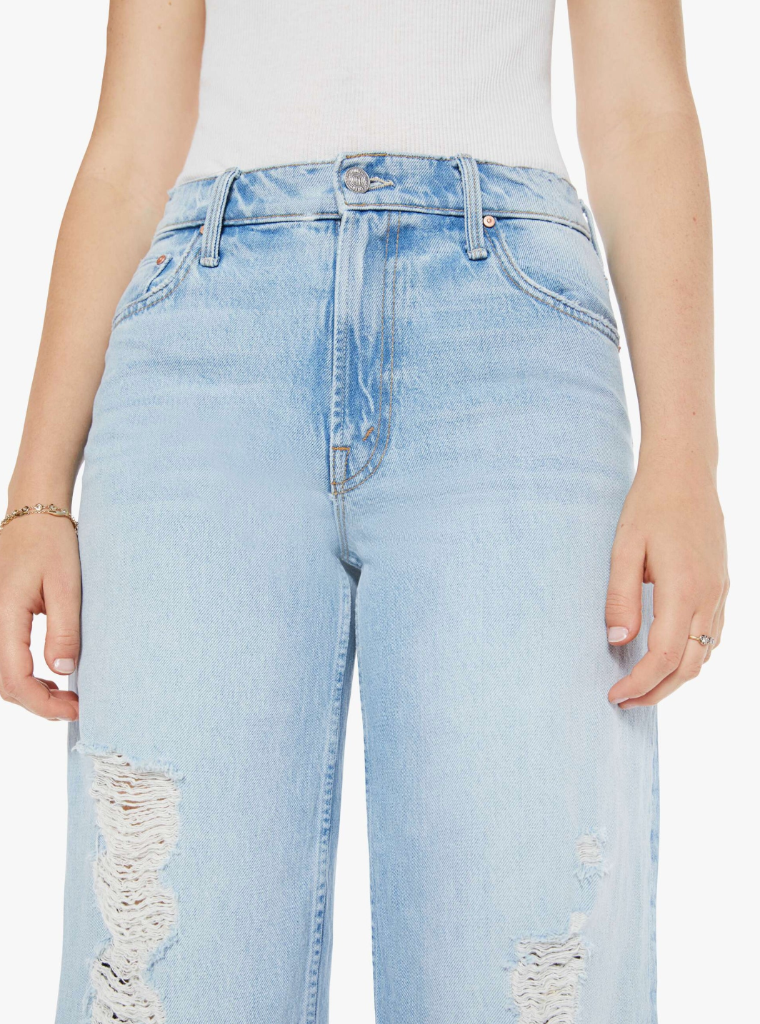 Wearing light blue high-waisted "The Spinner Zip Sneak" jeans by Mother, crafted from superior denim with distressed upper thigh details, they pair them with a white sleeveless top, hands casually at their sides.