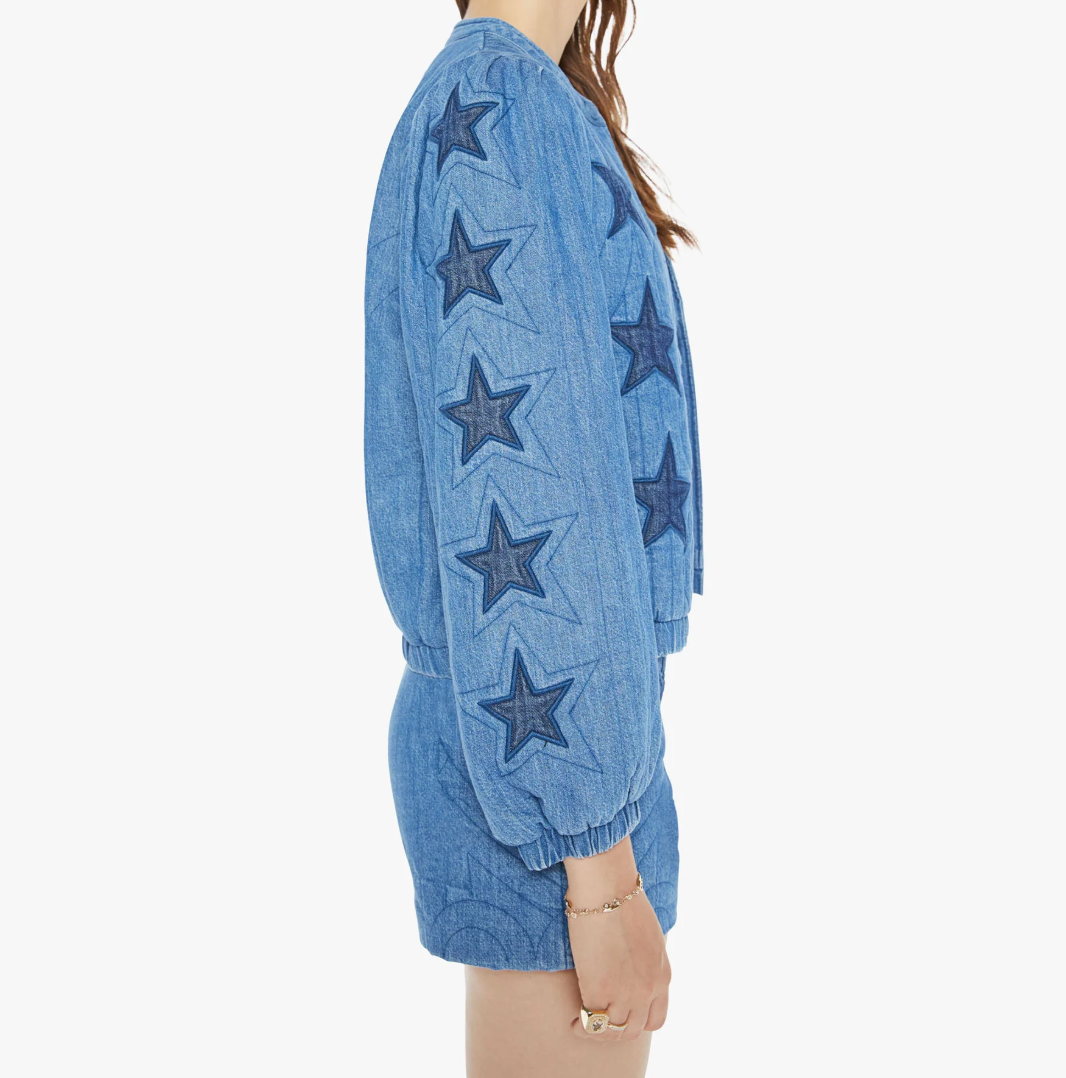 Wearing "The Rough Puff" by Mother, a chic blue denim jacket with star-patterned sleeves and quilted details, is stylishly paired with matching shorts against a plain white background. A side view accentuates the outfit's trendy elements.