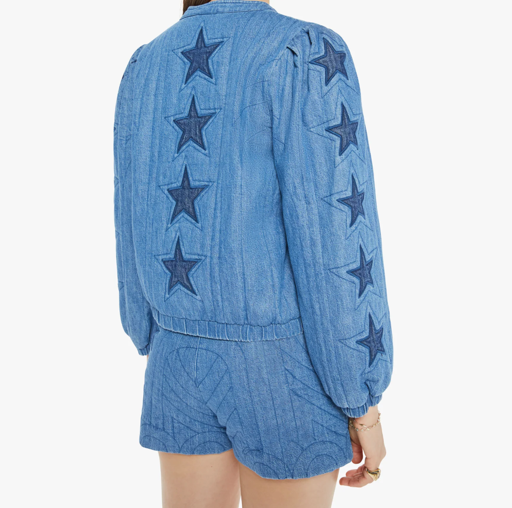 A person models "The Rough Puff" by Mother—a blue denim jacket with dark blue star patterns on the sleeves and back, echoing a quilted crew neck style. They pair it with matching wavy-textured shorts, all against a white backdrop.