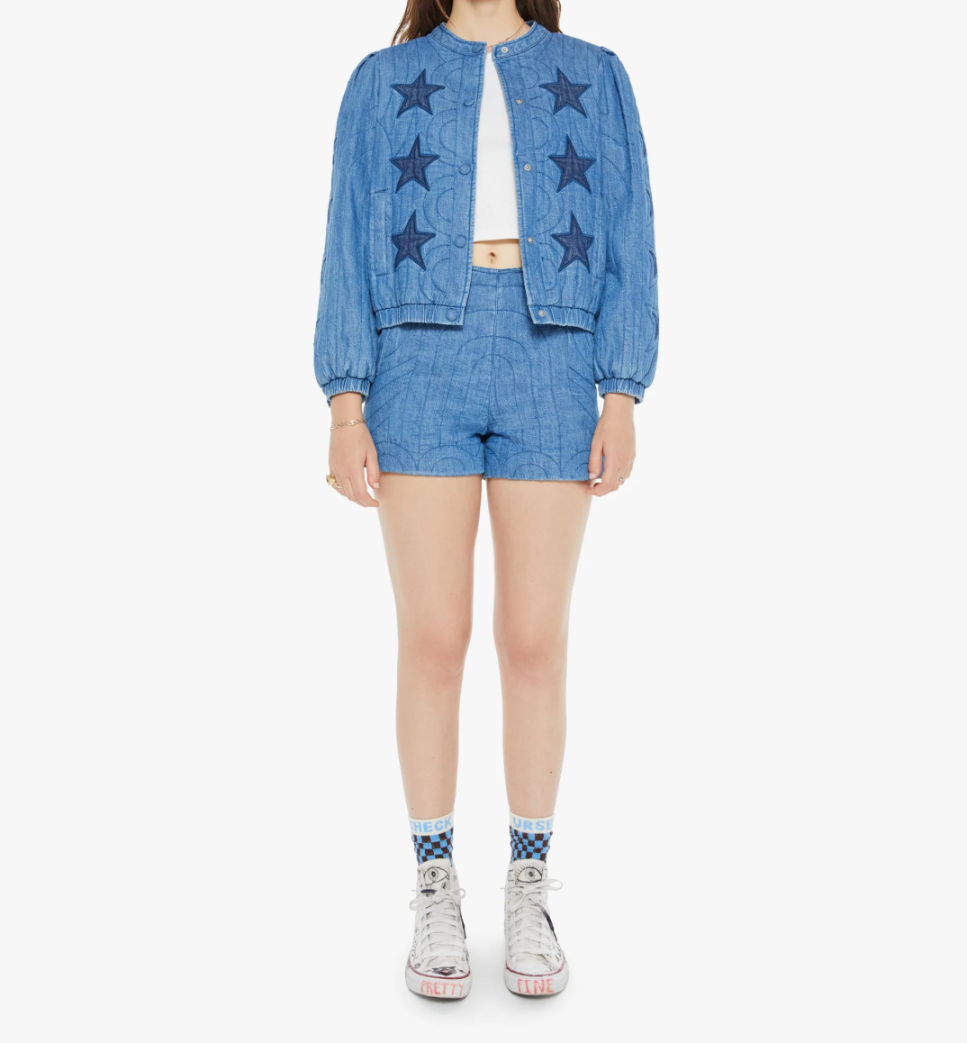 A person stands wearing "The Rough Puff" by Mother, a blue denim jacket with quilted details and star patterns, matching shorts, a white crop top, and white high-top sneakers. Their socks feature a geometric blue and white design. The background is plain white.