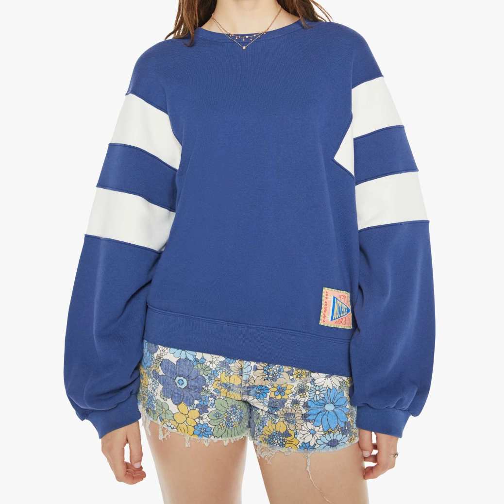 A person wears The Drop Square Inset, a blue, 100% cotton crewneck with two white sleeve stripes and a colorful bottom patch. They also sport Mother floral-patterned shorts in blue, yellow, and white.