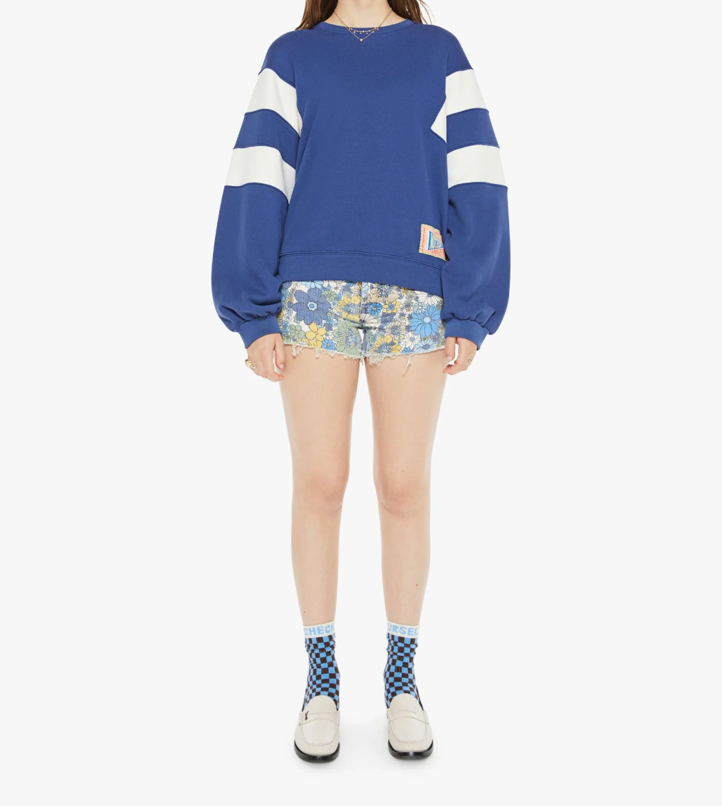 A person wearing an oversized blue Mother sweatshirt with white-striped sleeves, floral-patterned shorts, blue and white checkered socks, and white loafers stands against a pristine white background.