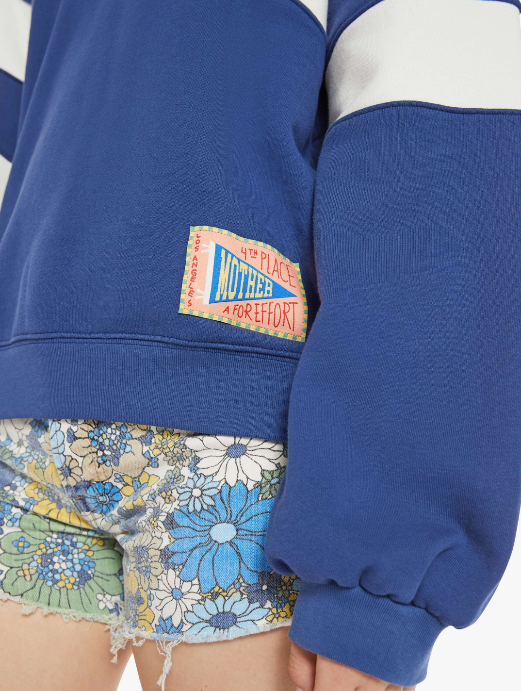 A person wears The Drop Square Inset, an oversized blue 100% cotton sweatshirt by Mother, featuring a "4th Place for Effort" patch and stylish drop shoulders. Their look is completed with vibrant blue, green, and yellow floral-patterned shorts.