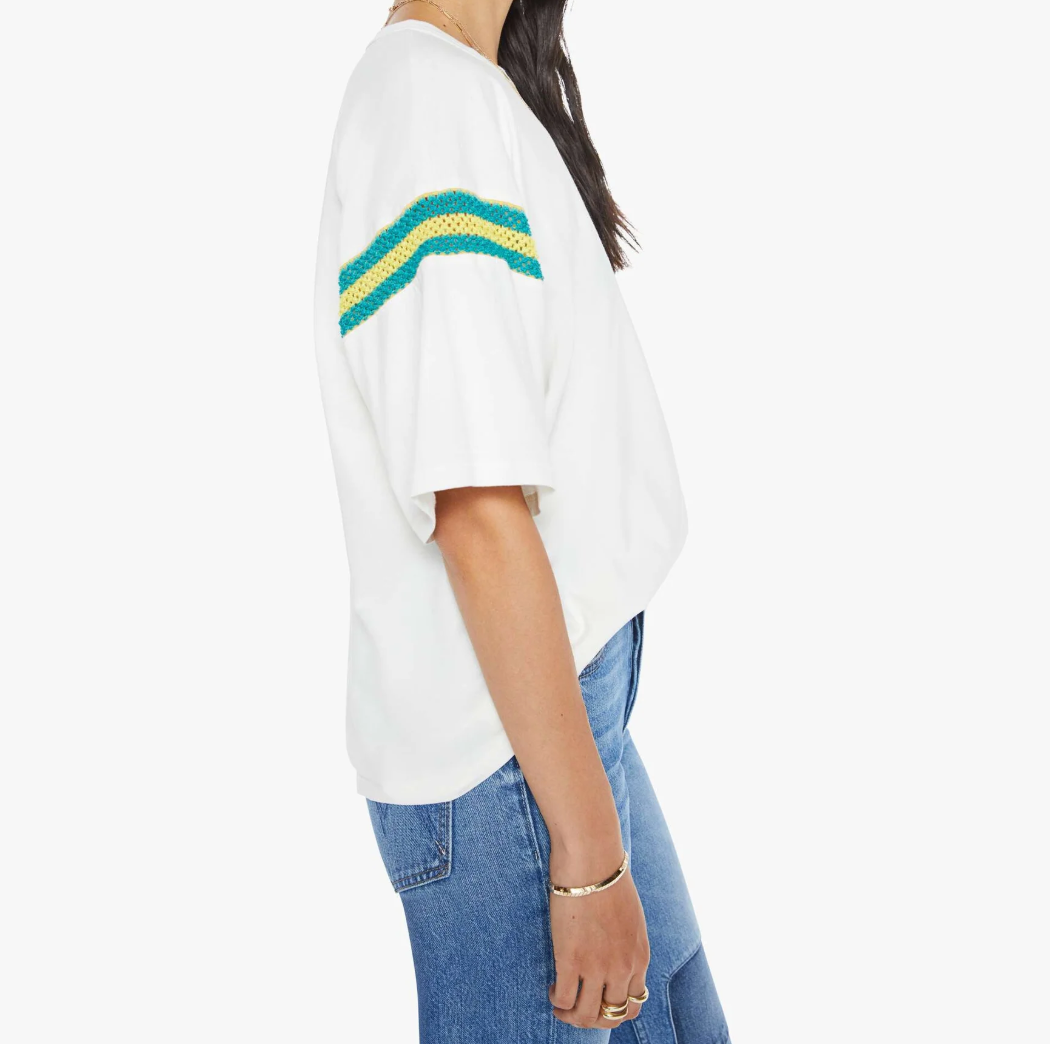 The person sports The Sleep Over Inset by Mother, a 100% cotton loose white T-shirt featuring green and yellow sleeve stripes with blue jeans and bracelets. Their head is slightly turned from the camera against an isolated white background, capturing a relaxed sleep-over vibe.