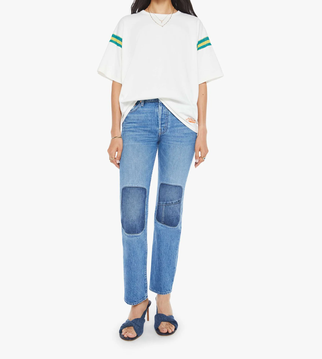 Dressed in The Sleep Over Inset by Mother, featuring an oversized white T-shirt with green striped sleeves paired with patched-knee blue denim jeans and chic blue heeled sandals, a person poses against a plain white background.