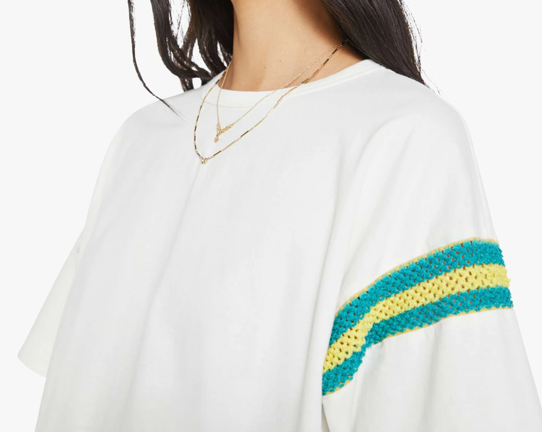 The woman wears Mother's "The Sleep Over Inset," an oversized white cotton T-shirt with teal and yellow crocheted sleeve stripes. Her long dark hair and layered gold necklaces perfectly complement the cozy sleep-over vibe, emphasizing her upper body and apparel details.