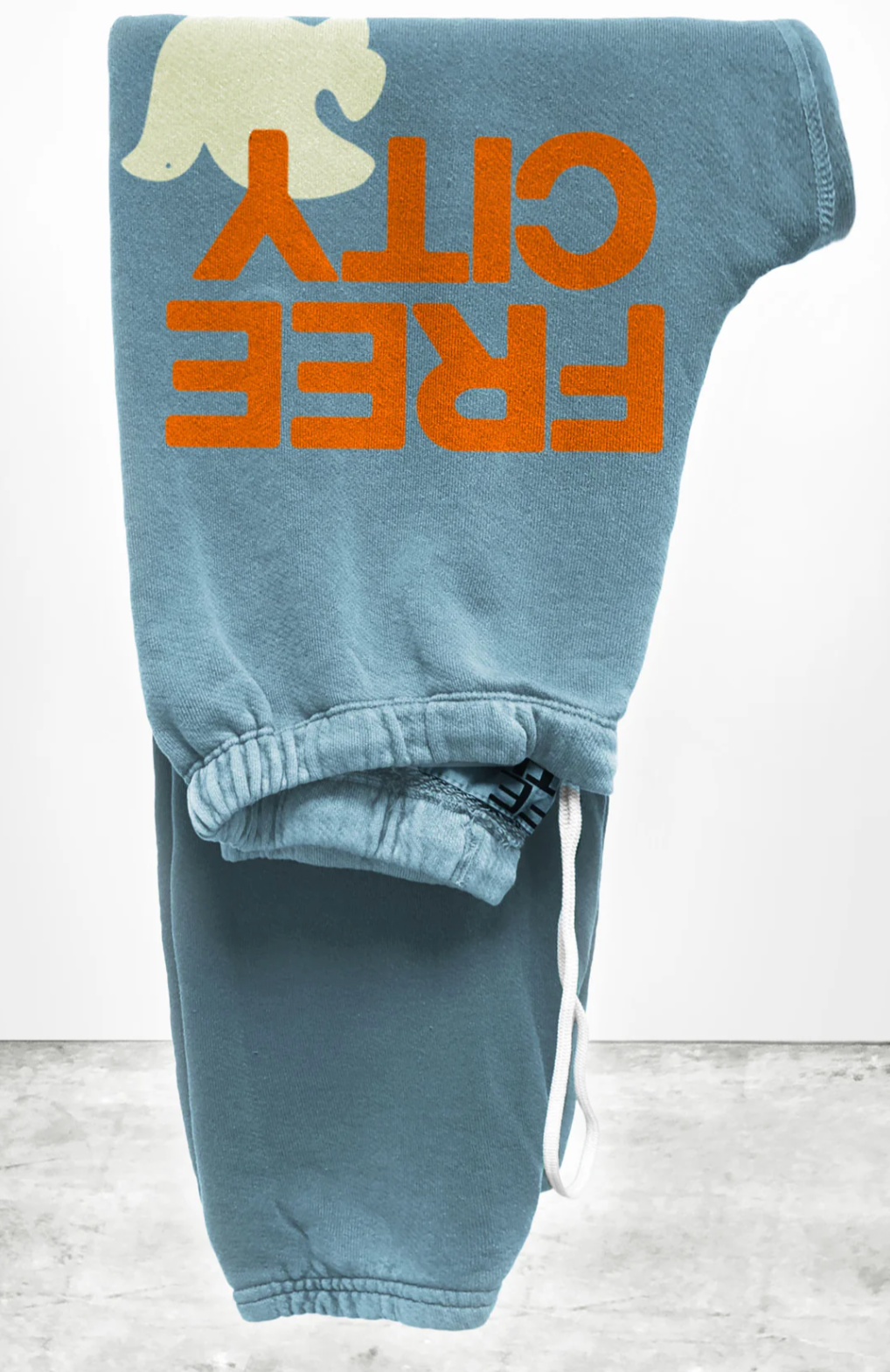 Folded blue FREECITY Large Sweatpant by Free City (sparrow, LLC), made from lightweight French terry, features an orange "FREE CITY" print and partial white graphic at the top left. Includes a white drawstring, placed on textured concrete against a plain background.