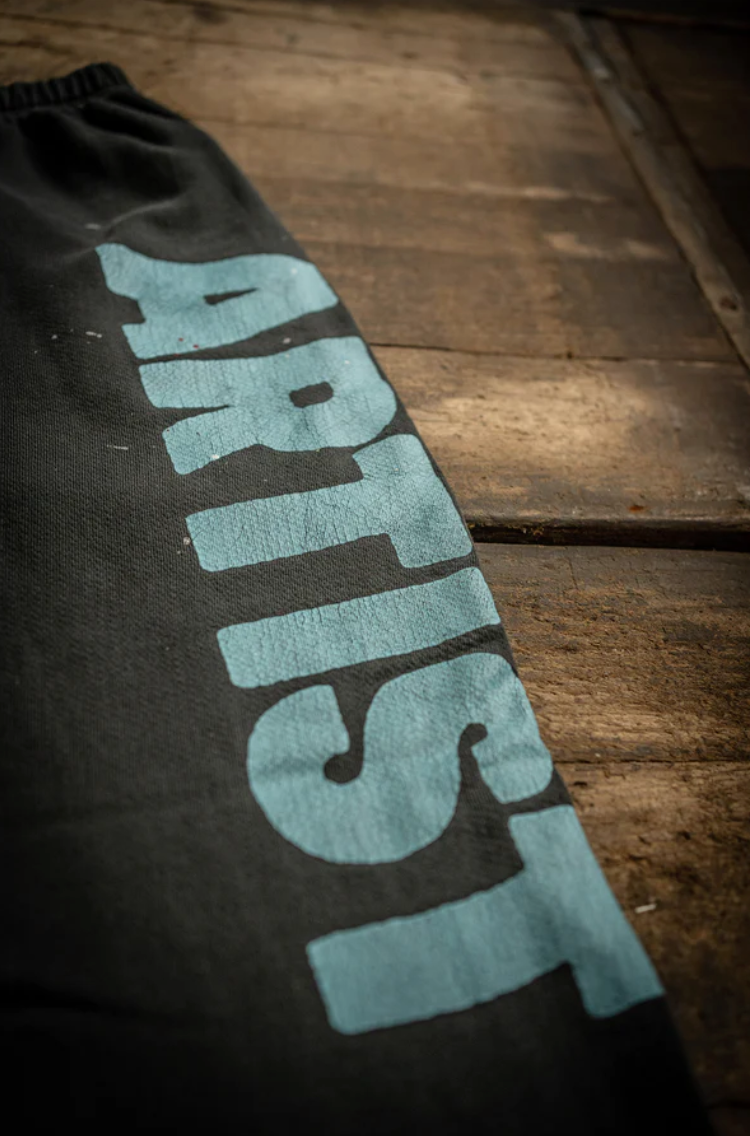 A close-up shows MadeWorn P.E. Sweatpants, a black French terry cotton fabric with "ARTIST" in large blue capital letters, offering a vintage unisex fit as it rests on a rustic wooden surface.