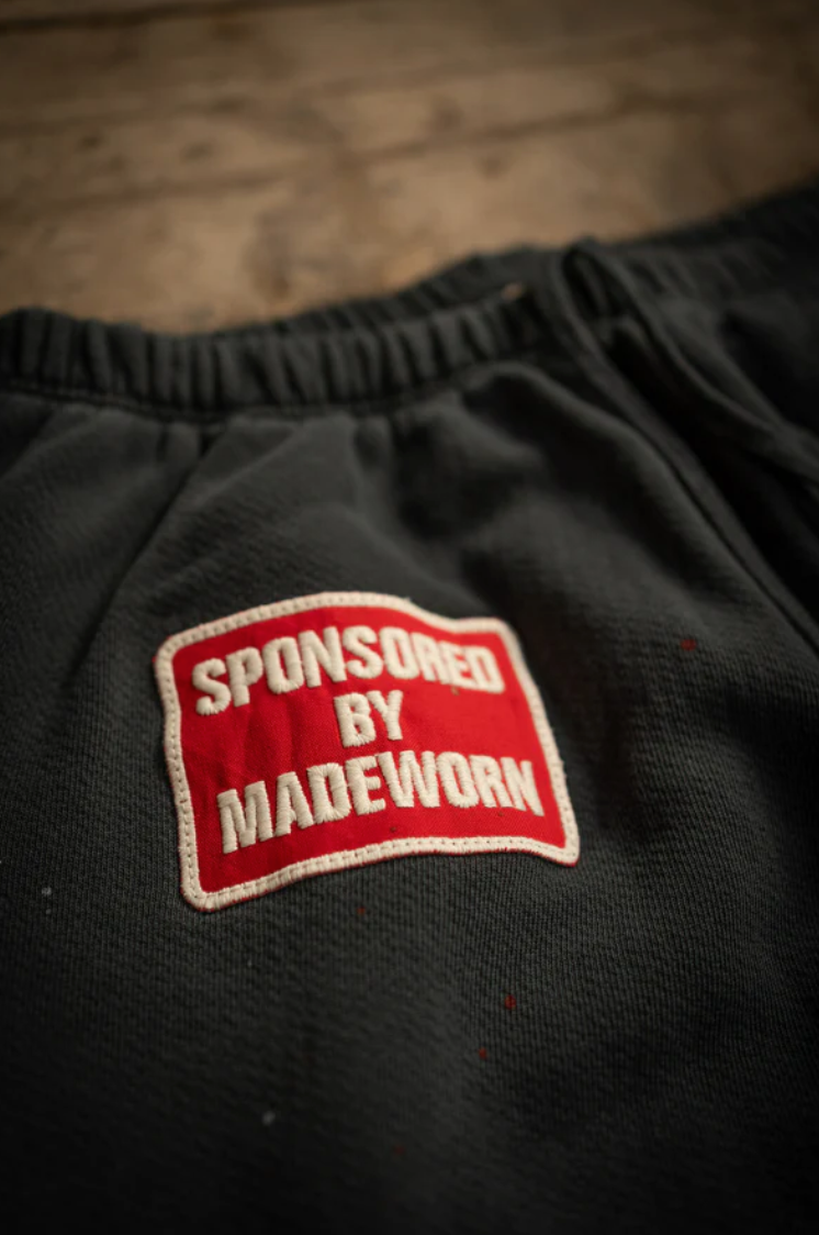 A close-up of dark fabric shows a stitched patch with "SPONSORED BY MADEWORN" in white on red. The heavyweight French terry cotton implies these could be MadeWorn P.E. Sweatpants, meticulously handmade in Los Angeles and potentially unisex.