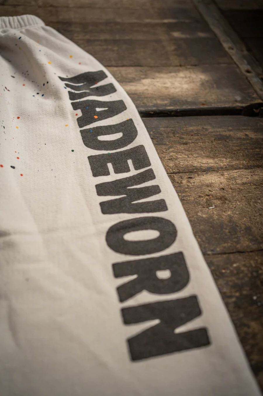 A close-up showcases a white fabric bag with "MADEWORN" in bold black letters, playfully splattered with colorful paint. This USA-made piece, displayed on rustic wood, evokes the vintage charm of PE Sweatpants by Made Worn crafted from French terry cotton.