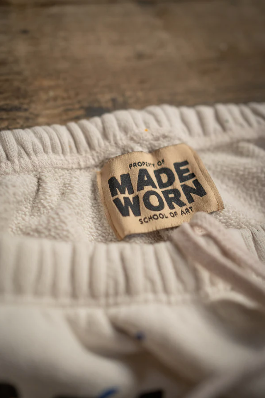 Close-up of the "PROPERTY OF MADE WORN SCHOOL OF ART" label on light-colored PE Sweatpant, crafted from French Terry Cotton with an elastic waistband. Displayed elegantly on a wooden surface, this Made Worn piece highlights its Made in USA craftsmanship.