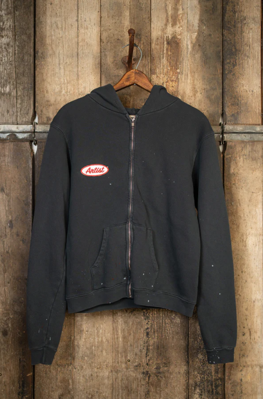 The MadeWorn Artist Hoodie, a black heavyweight French terry cotton zip-up, hangs on a rustic wooden wall with metal details. It features a unisex vintage fit and has a small red and white "Artists" patch on the left chest, showcasing its charm.