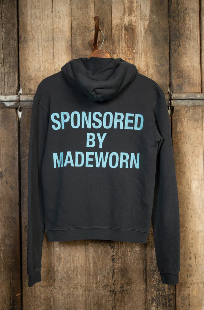 A MadeWorn Artist Hoodie in black hangs against a wooden backdrop, featuring large light blue text: "SPONSORED BY MADEWORN." This vintage unisex hoodie is handcrafted in Los Angeles from heavyweight French terry cotton.