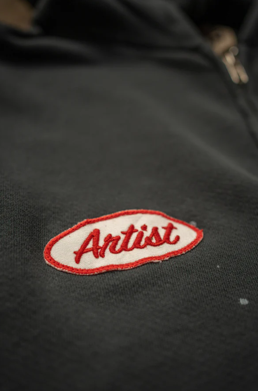 Close-up of the MadeWorn Artist Hoodie by Made Worn, a black vintage unisex design crafted from French terry cotton. It showcases a red and white embroidered oval patch with "Artist" in red cursive, highlighting quality craftsmanship with a small visible zipper, made in the USA.