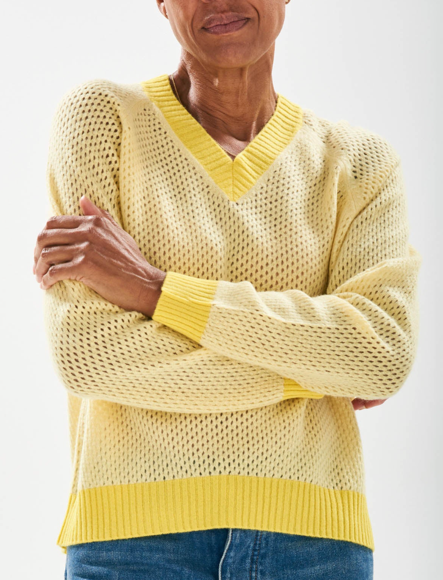 Wearing the Holey Contrast Vee by Jumper 1 2 3 4/CR2, a person in an oversized light yellow knit sweater with a V-neck and white contrast ribs stands with arms crossed. Paired seamlessly with blue jeans against a white backdrop, it exudes effortless style and comfort.