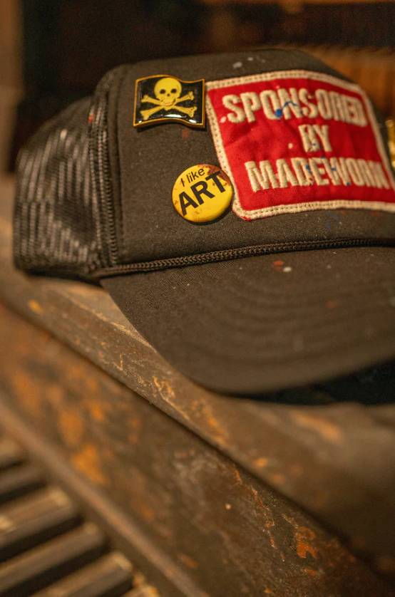 A limited edition black MadeWorn Sponsor Hat by Made Worn, featuring a skull and crossbones patch with "SPONSORED BY MADEUROWN" in red, rests on a worn wooden surface. A yellow pin states "I like ART," and an adjustable back strap enhances comfort. Piano keys are partially visible below.