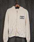 The MadeWorn Artist Hoodie, a white cotton zip-up, features a "Sponsored by MadeWorn" patch on the left chest and vibrant paint splatter detailing at the bottom. This handmade Los Angeles vintage unisex fit is elegantly displayed against a dark wooden wall.