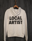 A white MadeWorn Artist Hoodie by Made Worn, crafted in Los Angeles, features "LOCAL ARTIST" on the back. Colorful paint splashes decorate the bottom and sleeves, enhancing its vintage unisex fit, as it hangs on a wooden wall.
