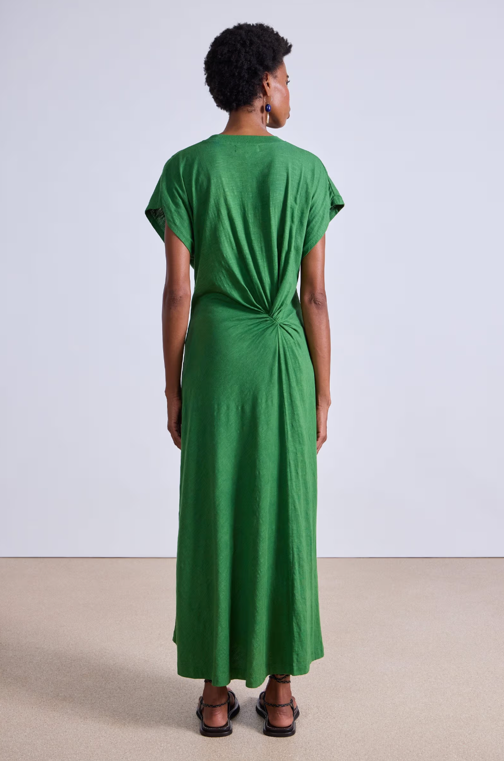 A person with short curly hair faces away, wearing Apiece Apart's Volta Twist Dress in long green organic cotton with a knotted back detail. They pair it with black sandals against a plain backdrop of a beige floor and light gray wall.
