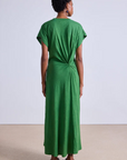 A person with short curly hair faces away, wearing Apiece Apart's Volta Twist Dress in long green organic cotton with a knotted back detail. They pair it with black sandals against a plain backdrop of a beige floor and light gray wall.