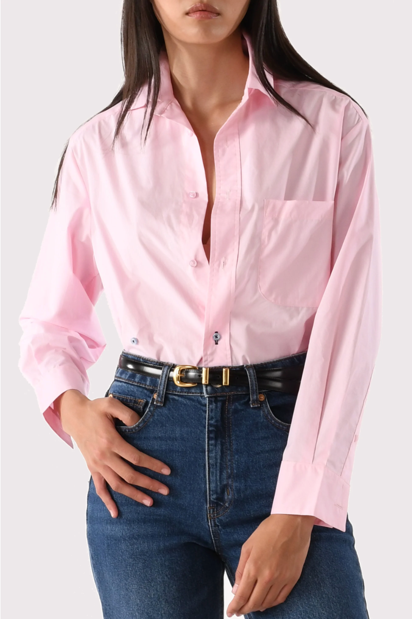 A person wears Cissa’s The Daily Shirt in light pink PAPER COTTON, tucked into high-waisted relaxed blue jeans with a black belt. With one hand on their hip, their face is partially out of the frame.