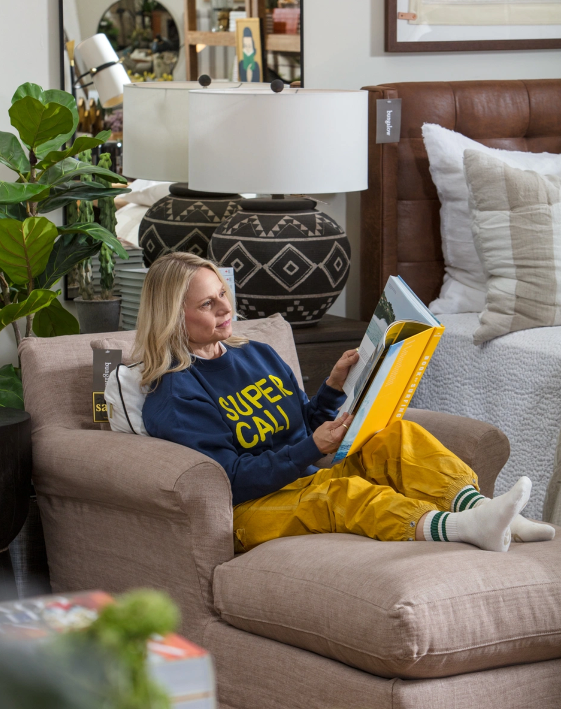 A woman in a Hype Factory Inc. SUPER CALI sweatshirt and yellow pants relaxes barefoot on a beige armchair, reading in a cozy, stylish living room filled with plants and decorative pillows.