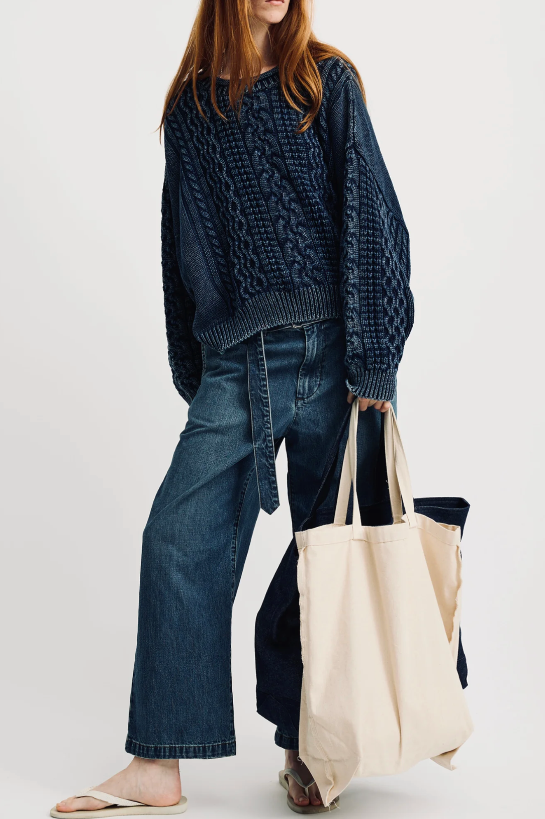 A person in the Denimist OVERSIZED ARAN SWEATER W/ROLLED NECK, wide-leg blue jeans, and sandals carries a large cream tote bag against a plain backdrop. Only their lower face and body are seen.
