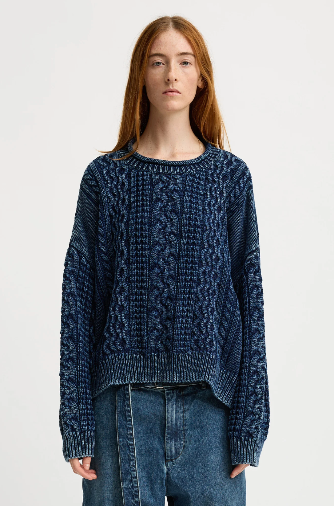 A person with long red hair wears the Denimist OVERSIZED ARAN SWEATER W/ROLLED NECK in navy blue with relaxed denim jeans, standing against a plain white background.