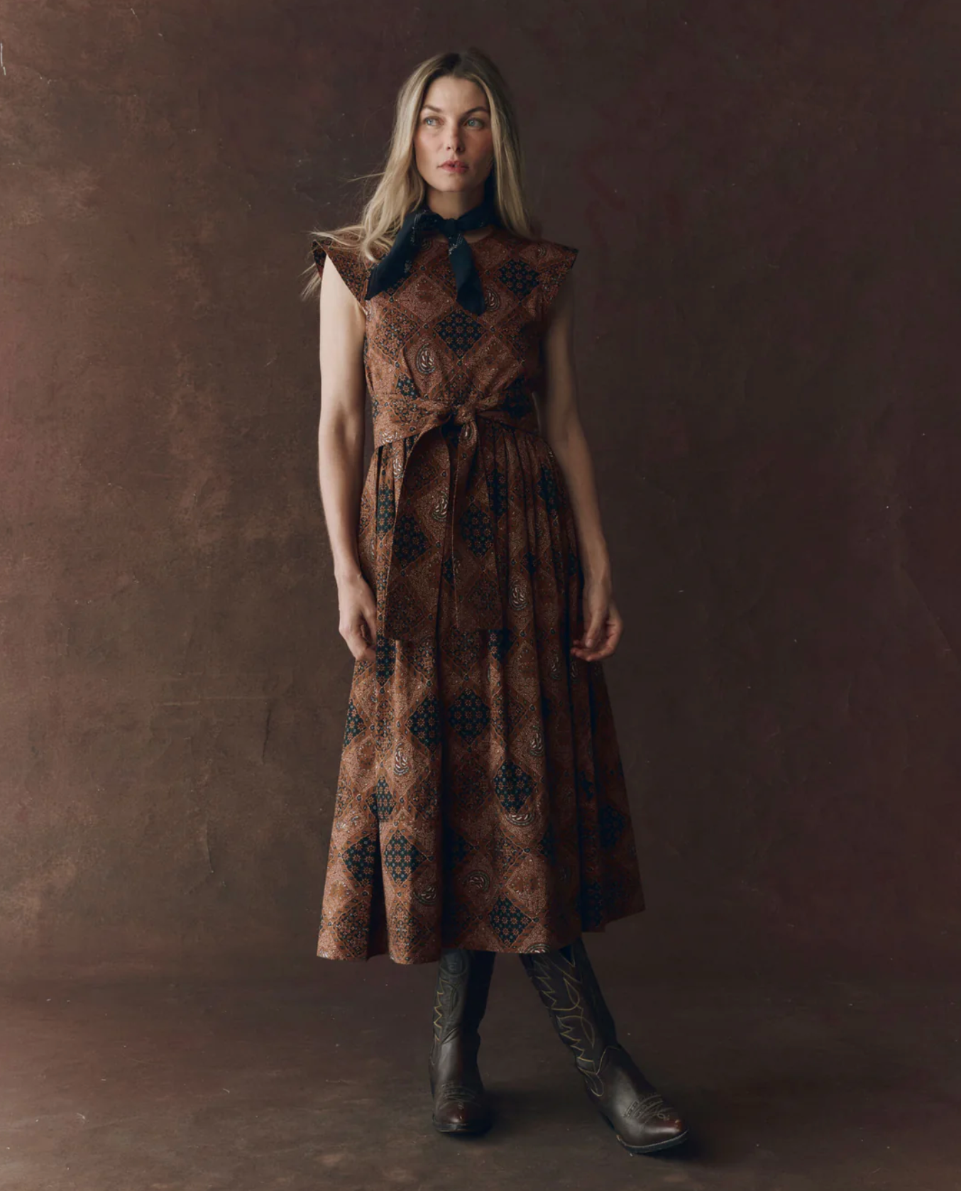 A woman with long light brown hair and a neutral expression stands before a dark brown backdrop, wearing The Great Inc.&#39;s Tulsa Dress—a brown patterned sleeveless dress with a waist seam, black necktie, and tall lace-up boots.