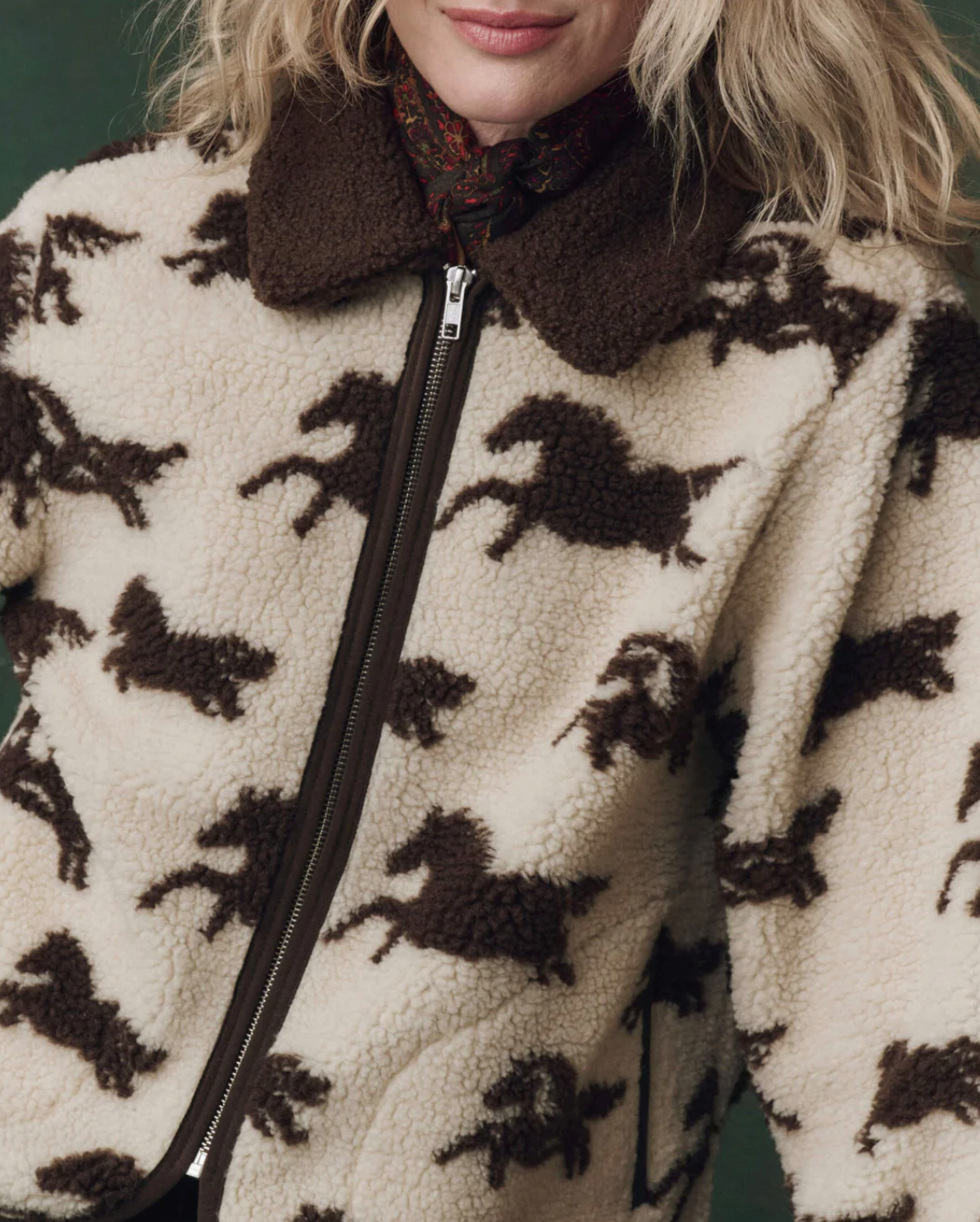 Someone wears The Pasture Jacket by The Great Inc.—a cream and brown relaxed-fit teddy jacket with a horse motif, brown collar, and visible zipper. The person’s face is partially out of frame.