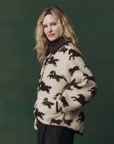 A person with long blonde hair wears The Great Inc.'s The Pasture Jacket, a cozy cream teddy coat featuring a black horse motif, standing against a dark green background and smiling slightly at the camera.