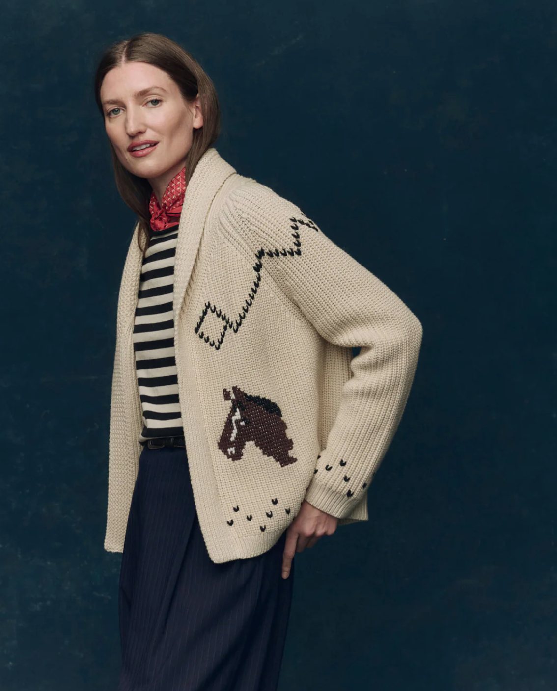 A person with long brown hair wears The Horse Lodge Cardigan by The Great Inc., featuring hand-embroidered horses, paired with a striped shirt, dark pleated pants, and a red patterned neck scarf against a dark blue background.