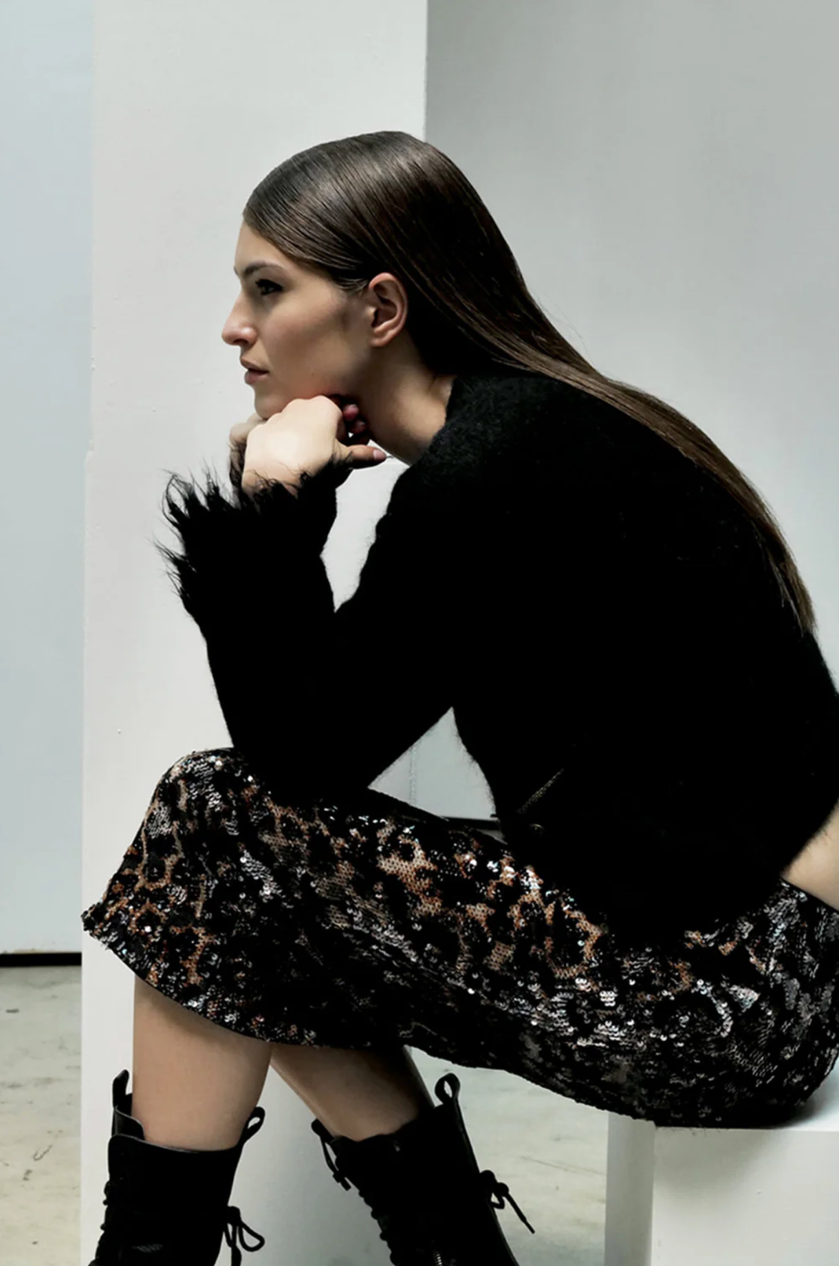 A woman with long, straight brown hair sits sideways on a white bench, resting her chin on her hand. She wears the Liza Skirt by Le Superbe with a black fuzzy sweater and black lace-up boots against a minimal white background.
