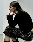 A woman with long, straight brown hair sits sideways on a white bench, resting her chin on her hand. She wears the Liza Skirt by Le Superbe with a black fuzzy sweater and black lace-up boots against a minimal white background.